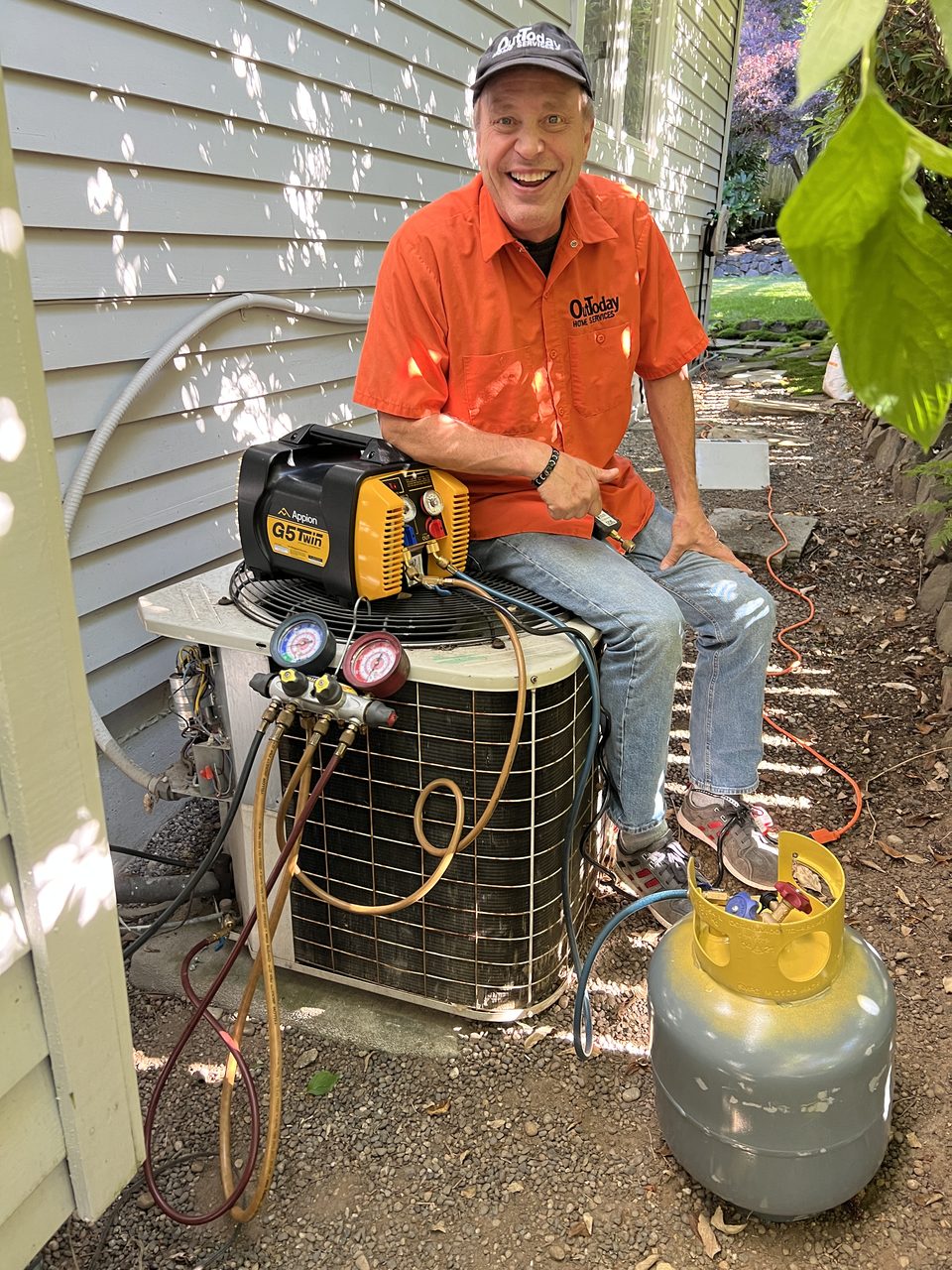 OutToday technician smiling while servicing an outdoor air conditioner with a refrigerant recovery machine and gauges